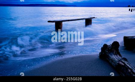 Eine ruhige Strandszene in der Abenddämmerung mit ruhigen Wellen, die sanft über eine provisorische Holzbank im Wasser fließen, Norwegian Point, WA, USA Stockfoto