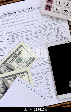 Zusammensetzung der Gegenstände, die auf dem Steuerformular 1040 liegen. Dollarscheine, Taschenrechner, Smartphone, Büroklammer und Notizblock. Die Zeit, Steuern zu zahlen Stockfoto