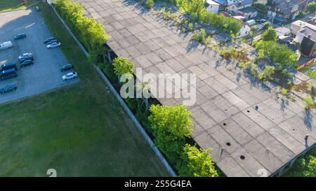 Blick aus der Vogelperspektive auf ein verlassenes Gebäude mit bewachsener Vegetation auf dem Dach, umgeben von einem Parkplatz und Wohngebiet. Stockfoto