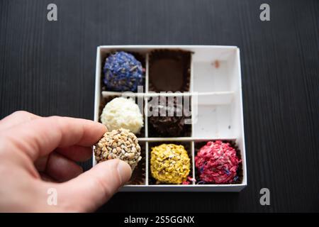 Eine Hand hält eine mit Nüssen überzogene Trüffel aus einer Schachtel mit verschiedenen bunten Trüffeln. Die Schachtel enthält verschiedene Trüffel mit verschiedenen Beschichtungen, einschließlich Stockfoto