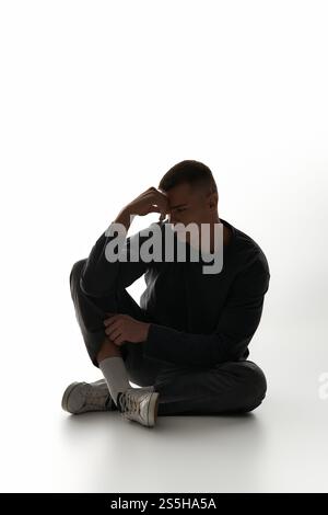 Ein junger Mann sitzt nachdenklich, Hand auf Stirn, verloren in einem minimalistischen Raum. Stockfoto