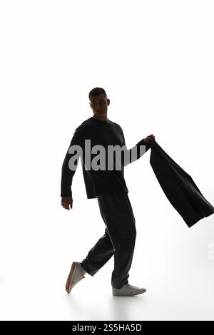 Ein junger Mann geht selbstbewusst mit einer Jacke vor einem hellen, minimalistischen Hintergrund. Stockfoto