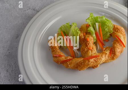 Frisch gebackene weiche Brezel mit großzügigem Streusel von grobem Salz und Sesam auf weißem Geschirr, mit Salat und Karotte schön dekorieren, Kopierraum Stockfoto
