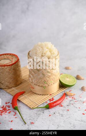 Thailändischer Kleberreis in einem gewebten Bambuskorb auf einer Holzplatte mit Chilis, Limette und Knoblauch. Selektiver Fokus. Stockfoto