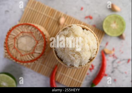 Thailändischer Kleberreis in einem gewebten Bambuskorb auf einer Holzplatte mit Chilis, Limette und Knoblauch. Draufsicht. Stockfoto