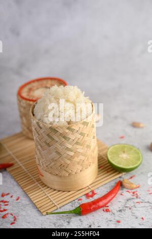 Thailändischer Kleberreis in einem gewebten Bambuskorb auf einer Holzplatte mit Chilis, Limette und Knoblauch. Selektiver Fokus. Stockfoto
