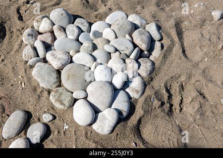 Eine Herzform aus glatten Kieselsteinen liegt auf dem Sand am Strand und zeigt einen künstlerischen Touch in einer natürlichen Umgebung. Stockfoto