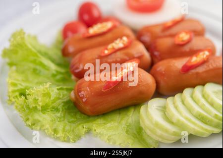 Die Wurst wird auf einem weißen Teller mit Chili, Tomaten, Gurken und Salatgemüse gelegt. Stockfoto