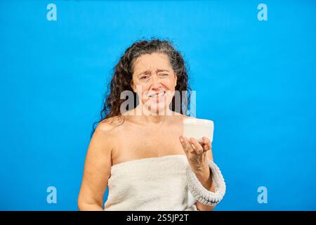 Frau mittleren Alters, die in ein Handtuch gewickelt ist und Ekel ausdrückt, während sie Kosmetikcreme hält, isoliert auf blauem Hintergrund Stockfoto