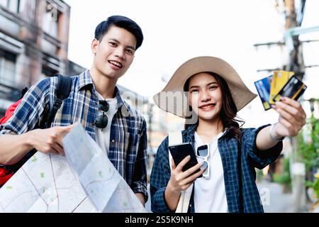 Ein junger Rucksackreisender, der eine Karte in Papierform hält, und eine hübsche Frau trägt einen Sombrero, hält ein Smartphone und zeigt eine Kreditkarte in der Hand, sie benutzen sie, um zu bezahlen Stockfoto