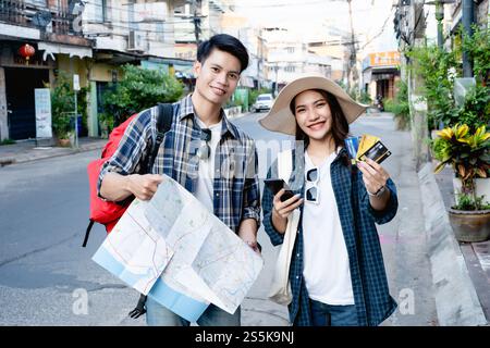 Ein junger Rucksackreisender, der eine Karte in Papierform hält, und eine hübsche Frau trägt einen Sombrero, hält ein Smartphone und zeigt eine Kreditkarte in der Hand, sie benutzen sie, um zu bezahlen Stockfoto