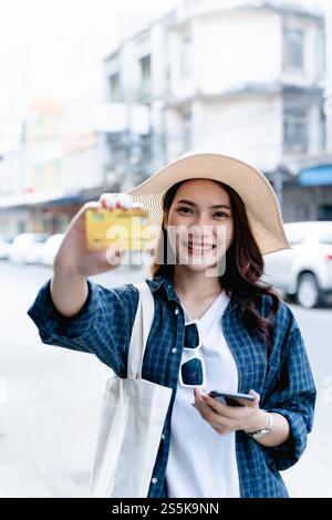 Selektiver Fokus junge Backpackerin weiblich im Sombrero lächelnd, während sie Smartphone hält und Kreditkarte in den Händen hält Stockfoto