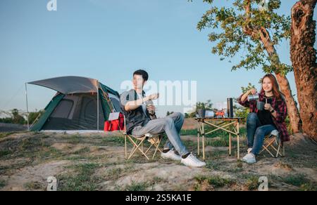 Fröhliches junges Backpacker-Paar, das vor dem Zelt im Wald mit Kaffee-Set sitzt und während eines Campingausflugs im Sommer frische Kaffeemühle zubereitete Stockfoto
