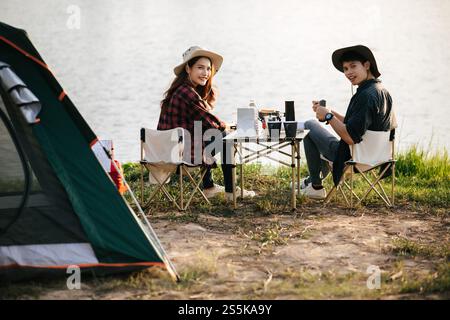 Fröhliches junges Backpacker-Paar, das vor dem Zelt im Wald mit Kaffee-Set sitzt und während eines Campingausflugs im Sommer frische Kaffeemühle zubereitete Stockfoto
