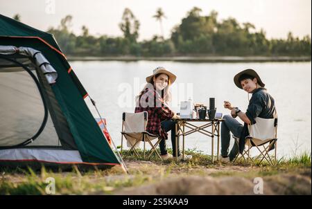 Fröhliches junges Backpacker-Paar, das vor dem Zelt im Wald mit Kaffee-Set sitzt und während eines Campingausflugs im Sommer frische Kaffeemühle zubereitete Stockfoto