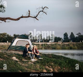 Fröhliches junges Backpacker-Paar, das vor dem Zelt in der Nähe des Sees mit Kaffee-Set sitzt und während des Campingausflugs im Sommer frische Kaffeemühle zubereitete Stockfoto