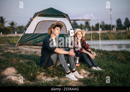Fröhliches junges Backpacker-Paar, das vor dem Zelt in der Nähe des Sees mit Kaffee-Set sitzt und während des Campingausflugs im Sommer frische Kaffeemühle zubereitete Stockfoto