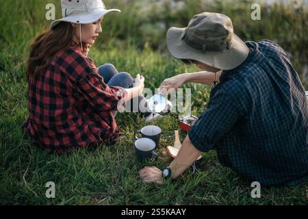 Fröhliches junges Backpacker-Paar, das auf Gras sitzt und sich bei heißem Kaffee während eines Campingausflugs im Sommerurlaub entspannt Stockfoto