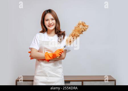 Porträt glückliche junge hübsche Frau in Schürze und Gummihandschuhen, die einen Federstaub halten, sich auf die Reinigung vorbereiten, lächeln und zur Kamera schauen, Kopierraum Stockfoto