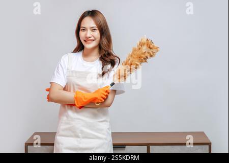 Porträt glückliche junge hübsche Frau in Schürze und Gummihandschuhen, die einen Federstaub halten, sich auf die Reinigung vorbereiten, lächeln und zur Kamera schauen, Kopierraum Stockfoto