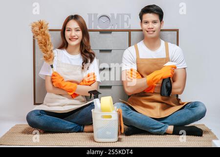 Porträt glückliches, lächelndes junges Paar mit Ausrüstung für das Putzhaus, sie tragen Gummihandschuhe, hübsche Frau hält Federstaub in der Hand, sie Stockfoto