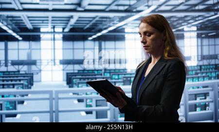 Akribischer Techniker, der die Überprüfung durchführt und Daten auf ein Tablet mit erforderlichen Fehlerbehebungen im Server-Hub eingibt, in dem eine fortschrittliche Datenspeicherinfrastruktur zur Unterstützung kritischer IT-Workloads, Kamera B, untergebracht ist Stockfoto