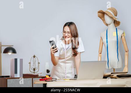 Schöne junge asiatische Modedesignerin oder Schneiderin, die mit Laptop-Computer arbeiten und Smartphone während der Arbeit verwenden, voll von Schneidewerkzeugen mit Stockfoto