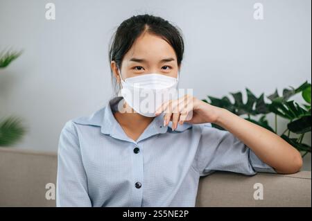 Junge asiatische Frau mit Gesichtsmaske, die auf dem Sofa im Wohnzimmer sitzt, sie liest Buch während der Quarantäne covid-19-Selbstisolierung zu Hause Stockfoto