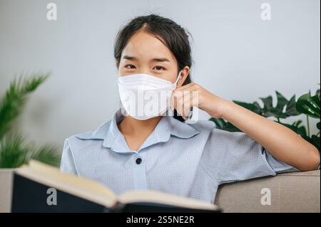 Junge asiatische Frau mit Gesichtsmaske, die auf dem Sofa im Wohnzimmer sitzt, sie liest Buch während der Quarantäne covid-19-Selbstisolierung zu Hause Stockfoto