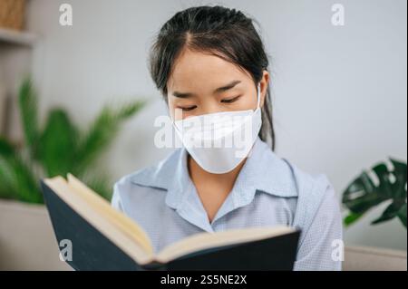 Junge asiatische Frau mit Gesichtsmaske, die auf dem Sofa im Wohnzimmer sitzt, sie liest Buch während der Quarantäne covid-19-Selbstisolierung zu Hause Stockfoto