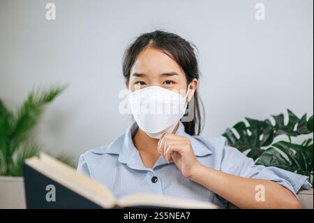 Junge asiatische Frau mit Gesichtsmaske, die auf dem Sofa im Wohnzimmer sitzt, sie liest Buch während der Quarantäne covid-19-Selbstisolierung zu Hause Stockfoto