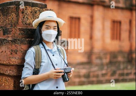 Junge asiatische Backpackerin, die Hut und Schutzmaske trägt, während sie durch die historische Stätte reist, benutzt sie Kamera, um ein Foto mit Happy zu machen Stockfoto
