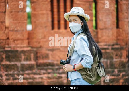 Junge asiatische Backpackerin, die Hut und Schutzmaske trägt, während sie durch die historische Stätte reist, benutzt sie Kamera, um ein Foto mit Happy zu machen Stockfoto