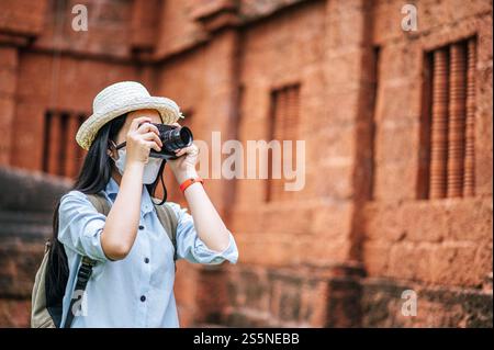 Junge asiatische Backpackerin, die Hut und Schutzmaske trägt, während sie durch die historische Stätte reist, benutzt sie Kamera, um ein Foto mit Happy zu machen Stockfoto