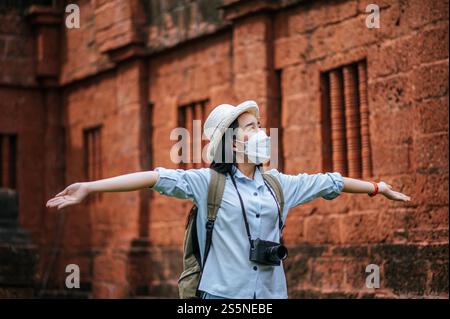 Junge asiatische Backpackerin, die Hut und Schutzmaske trägt, während sie durch die historische Stätte reist, benutzt sie Kamera, um ein Foto mit Happy zu machen Stockfoto