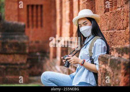 Junge asiatische Backpackerin, die Hut und Schutzmaske trägt, während sie durch die historische Stätte reist, benutzt sie Kamera, um ein Foto mit Happy zu machen Stockfoto