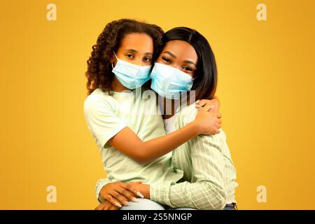 Porträt afroamerikanischer Mutter und Tochter, die medizinische Schutzmasken auf dem Gesicht auf gelbem Hintergrund tragen Stockfoto