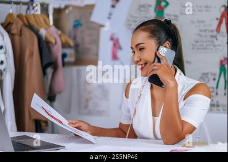 Die junge Modedesignerin, die mit dem Smartphone spricht und auf der Zeichnung der Kleidung schaut, lächelt mit Happy im Schneiderladen Stockfoto