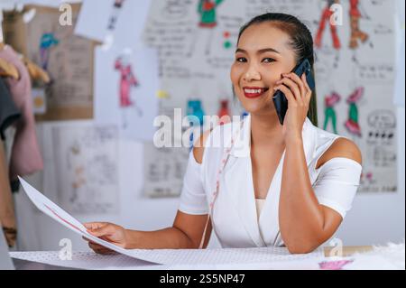 Die junge Modedesignerin, die mit dem Smartphone spricht und auf der Zeichnung der Kleidung schaut, lächelt mit Happy im Schneiderladen Stockfoto