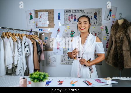 Porträt Junge asiatische Designerin, die in der Werkstatt steht, lächelt und die Kamera sieht, zeichnet Modeskizze auf dem Tisch. Maßband am Hals, Stockfoto