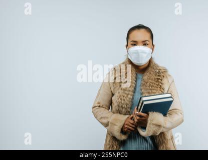 Porträt einer asiatischen Frau, die Bücher in der Hand hält und einen Pelzmantel trägt, zum Schutz vor Coronavirus Schutzmaske auf das Gesicht aufsetzt, stehend Stockfoto