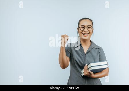Porträt einer asiatischen Frauenbrille, die Bücher in der Hand hält und Erfolg feiert, während sie die Kamera über dem Hintergrund im Studio mit Kopierraum betrachtet. Stockfoto