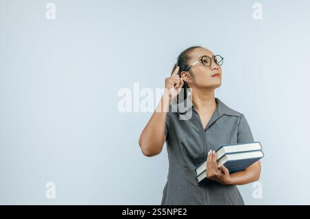 Porträt einer seriösen asiatischen Frauenbrille, die mit einem Finger auf den Kopf zeigt und Bücher in der Hand über Hintergrund im Studio mit Kopierraum hält. Stockfoto