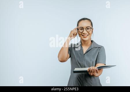 Porträt einer glücklichen asiatischen Frau Brille, die mit einem Finger auf den Kopf zeigt und Bücher in der Hand über Hintergrund im Studio mit Kopierraum hält. Weiblich Stockfoto