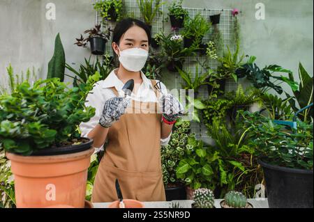 Asiatische Gärtner Frau trägt Gesichtsmaske und Schürze Stand und zeigt eine Schaufel und Heugabel, Ausrüstung, um Pflanzen in Innenräumen zu transplantieren und kümmern sich um Stockfoto