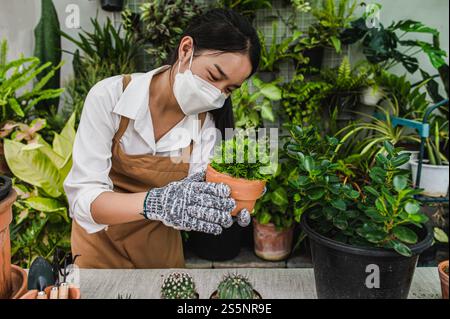 Asiatische Gärtner Frau trägt Gesichtsmaske und Schürze, die grüne Zimmerpflanze in der Hand hält, während sie sich um Innenhauspflanze zu Hause, Hobby oder Kleinunternehmen kümmern Stockfoto
