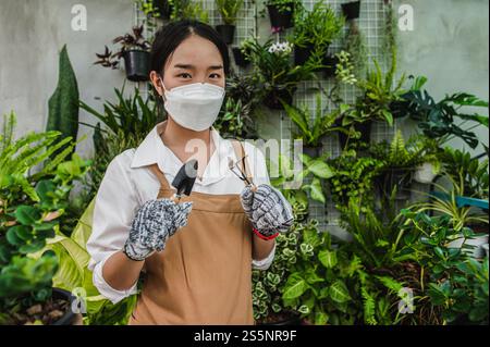 Asiatische Gärtner Frau trägt Gesichtsmaske und Schürze Stand und zeigt eine Schaufel und Heugabel, Ausrüstung, um Pflanzen in Innenräumen zu transplantieren und kümmern sich um Stockfoto