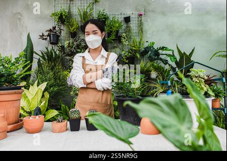 Asiatische Gärtnerin trägt Gesichtsmaske und Schürze mit Schaufel, um Kakteen und Zimmerpflanzen zu transplantieren und sich um Pflanzen zu Hause, Hobby oder klein zu kümmern Stockfoto