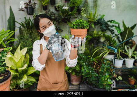 Asiatische Gärtner Frau trägt Gesichtsmaske und Schürze, die Spray zur Bewässerung der grünen Zimmerpflanze in der Hand hält, während sie sich um die Innenhauspflanze auf dem Tisch kümmern Stockfoto