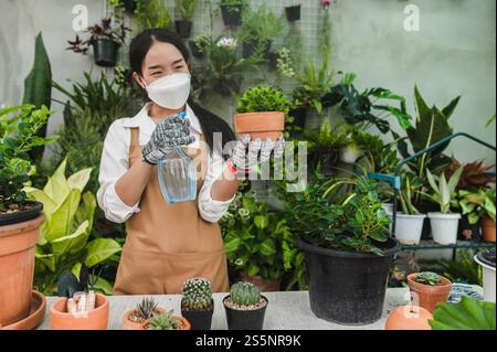 Asiatische Gärtner Frau trägt Gesichtsmaske und Schürze, die Spray zur Bewässerung der grünen Zimmerpflanze in der Hand hält, während sie sich um die Innenhauspflanze auf dem Tisch kümmern Stockfoto